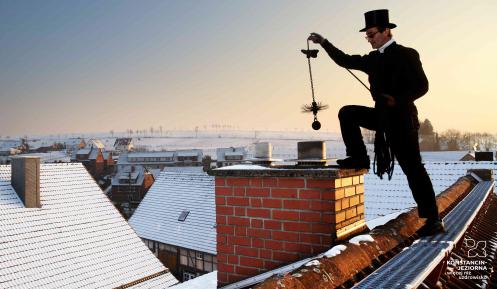 Mężczyzna, kominiarz stoi na dachu. Ubrany jest w czarną kurtkę, na głowie ma czarny kapelusz. Czysci komin. W tle domy jednorodzinne i błękitne niebo.