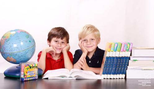 Dwóch chłopców (blondyn i szatyn) siedzi uśmiechniętych przy biurku. Na biurku znajdują się: globus, podręczniki, atlasy i duzy globus.
