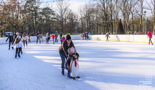 Park zdrojowy, ludzie w strojach zimowych jeżdżą na lodowisku, w tle budynek amfiteatru i drzewa.