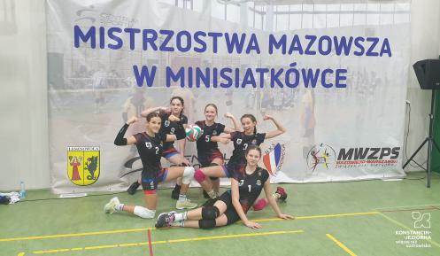 Pięć dziewcząt w czarnych strojach sportowych przyklęka na jednym kolanie i  pozuje do zdjęcia na tle baneru z napisem: mistrzostwa Mazowsza w minisiatkówce.