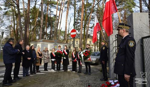 Grupa młodych dziewczyn i chłopców z biało-czerwonymi wieńcami i szarfami oraz trzech starszych mężczyzn i trzy kobiety stoją naprzeciwko tablicy zawieszonej na ogrodzeniu. Przy ogrodzeniu dwóch strażników miejskich w galowych mundurach oraz biało-czerwone flagi.