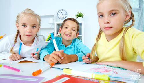 Dwie dziewczynki z blond warkoczykami, a w środku chłopiec. Siedzą za stołem w szkolnej klasie i rysują.