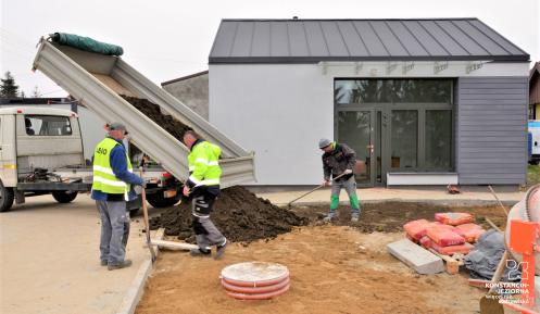 Budowa, wywrotka zrzuca ziemie, 3 robotników stoi obok. W tle widać szary budynek. 