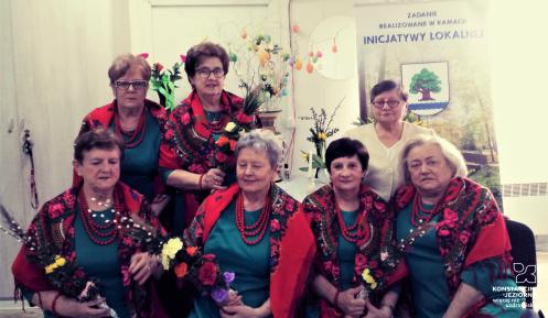 Siedem starszych kobiet w strojach ludowych, mają palmy wielkanocne w dłoniach. Za nimi baner z napisem: „Zadanie realizowane w ramach inicjatywy lokalnej”. 