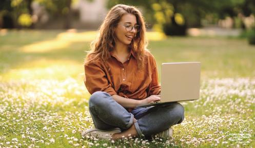 Uśmiechnięta dziewczyna siedzi na łące. Ma długie włosy i jest w okularach. Na kolanach ma otwarty laptop.