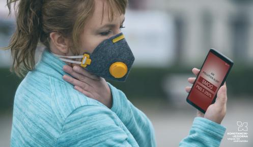 Kobieta z maseczką na twarzy patrzy w monitor telefonu, na który wyświetlony jest napis " Air pollution alert: Smog"