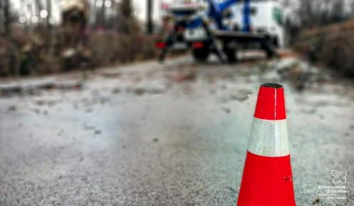 Czerwono-biały pachołek ostrzegawczy na ulicy. W tle widać drzewa i podnośnik do ich ścinania.