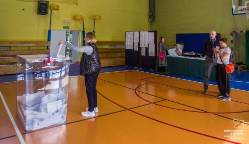 Pomieszczenie sali gimnastycznej, kobieta wrzuca kartkę do urny wyborczej 