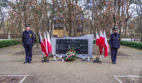 Pośrodku czarna tablica granitowa z tekstem umocowana na prostokątnym kamieniu, po lewej i prawej stronie stoją flagi narodowe, przed tablicą leżą kwiaty i wiązanki oraz palą się znicze