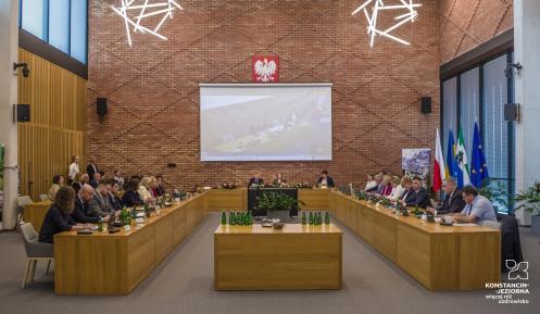 Grupa osób siedzi przy stołach w sali posiedzeń.