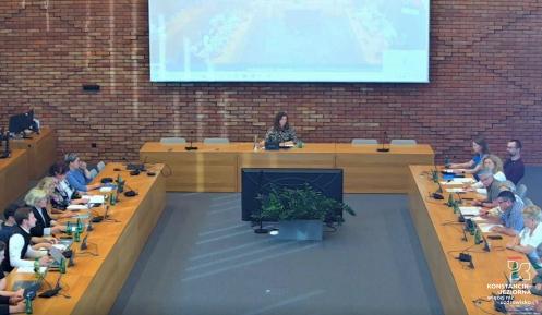 Sala posiedzeń, około 20 osób siedzi przy stołach, na ścianie wisi ekran projekcyjny