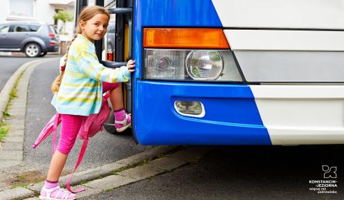 Dziewczynka ubrana w różową sukienkę i zielony sweter wsiada do biało-niebieskiego autobusu.