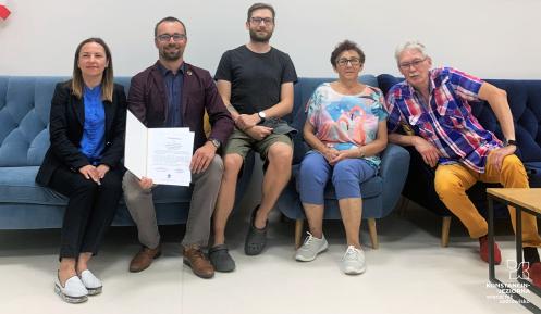 Grupa pięciu osób siedzi na kanapie, jedna z nich prezentuje dyplom