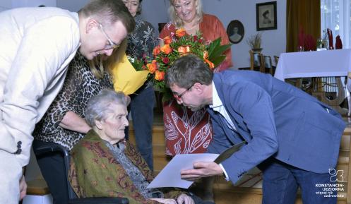 Mężczyzna z listem gratulacyjnym pochylony nad starszą kobietą, w tle inne osoby.