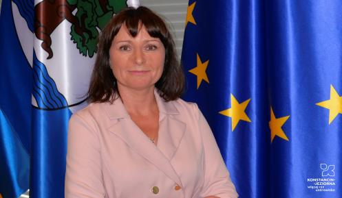 Kobieta z założonymi rękoma na tle flag Konstancina-Jeziorny i Unii Europejskiej.