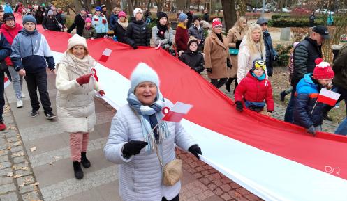 Grupa osób, w tym dzieci i osoby starsze, bierze udział w marszu z okazji Święta Niepodległości. Uczestnicy trzymają dużą, biało-czerwoną flagę Polski, a niektórzy mają przypięte kotyliony.