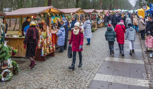 Na zdjęciu widać jarmark bożonarodzeniowy z rzędem kolorowych stoisk i spacerującymi ludźmi w zimowych ubraniach na brukowanej ścieżce wśród drzew.