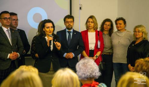 Grupa osób stoi na scenie, a kobieta w ciemnym żakiecie przemawia do mikrofonu przed publicznością.