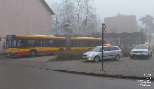 Żółto-czerwony autobus przegubowy warszawskiej komunikacji miejskiej zaparkowany na pętli autobusowej, w otoczeniu budynków i pawilonów handlowych spowitych mgłą. Obok stoją dwa pojazdy techniczne Zarządu Transportu Miejskiego z włączonymi światłami ostrzegawczymi.