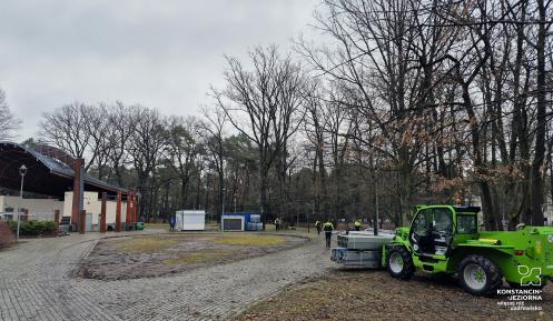 park z zadaszoną sceną, robotników w kamizelkach pracujących przy montażu lodowiska oraz zieloną ładowarkę teleskopową transportującą elementy konstrukcyjne.