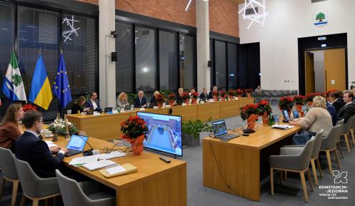 Zdjęcie przedstawia sesję Rady Miejskiej w nowoczesnej sali obrad, z radnymi przy stołach ozdobionych poinsecjami i flagami w tle.