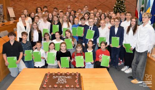 Grupa młodych sportowców trzymających zielone dyplomy pozuje do zdjęcia w sali konferencyjnej z dekoracyjną choinką i tortem z napisem Sportowiec Roku 2024 na pierwszym planie.
