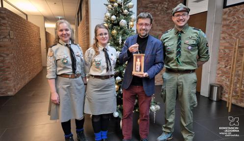 Grupa czterech osób stoi przed udekorowaną choinką, z czego trzy osoby są w mundurach harcerskich, a jedna trzyma zapaloną lampkę.