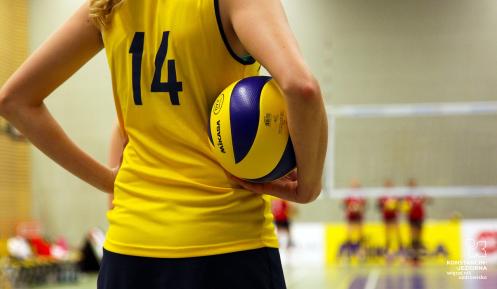 Zawodniczka siatkarska w żółtej koszulce z numerem 14 trzyma piłkę do siatkówki, stojąc na boisku przed meczem lub treningiem.