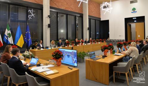 Zdjęcie przedstawia salę obrad z uczestnikami siedzącymi przy stołach, udekorowaną flagami Polski, Ukrainy, Unii Europejskiej oraz roślinami w świątecznym wystroju.