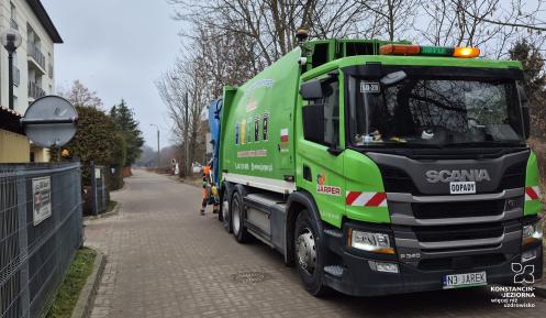 Zielona śmieciarka firmy Jarper marki Scania, stojąca na osiedlowej uliczce podczas odbioru odpadów przez pracownika w pomarańczowym stroju.
