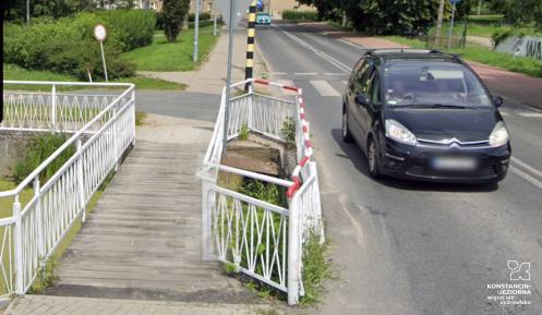 Widok na drewnianą kładkę przez rów wodny, obok samochód.
