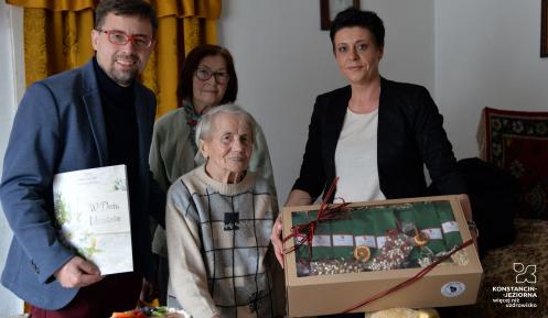 Grupa osób, w tym jubilatka, otrzymuje upominki i gratulacje z okazji setnych urodzin w domowym otoczeniu.