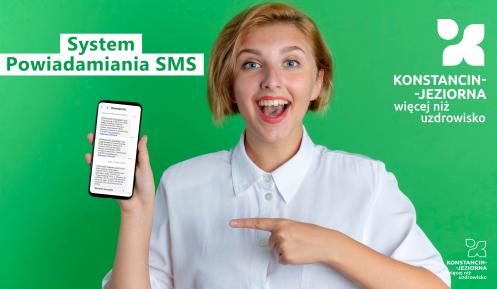 Młoda kobieta z szerokim uśmiechem trzyma w dłoni smartfon z wiadomościami SMS, na zielonym tle z napisami promującymi system powiadamiania SMS dla Konstancina-Jeziorny.