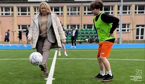 Kobieta w eleganckim stroju kopie piłkę na boisku sportowym, podczas gdy chłopiec w sportowym stroju obserwuje ją z uwagą.