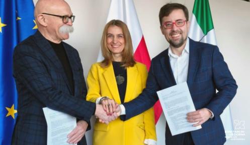 Trzech przedstawicieli podpisuje porozumienie, uśmiechając się i ściskając dłonie na tle flag Unii Europejskiej, Polski i Konstancina-Jeziorny.