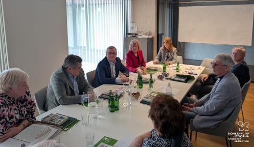 Grupa osób uczestniczy w formalnym spotkaniu przy stole konferencyjnym, omawiając dokumenty i notatki.