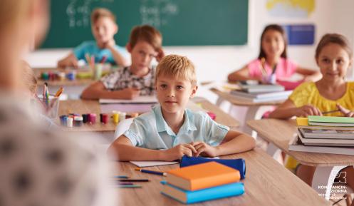 W klasie nauczycielka stoi przy tablicy, dzieci siedzą w ławkach i zgłaszają się do odpowiedzi