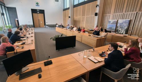 Spotkanie w nowoczesnej sali konferencyjnej, gdzie uczestnicy siedzą przy długich drewnianych stołach, wyposażonych w mikrofony i monitory, uważnie słuchając przemawiających osób.