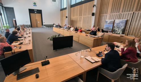 Spotkanie w nowoczesnej sali konferencyjnej, gdzie uczestnicy siedzą przy długich drewnianych stołach, wyposażonych w mikrofony i monitory, uważnie słuchając przemawiających osób.