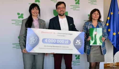 Trzy osoby pozują do zdjęcia, trzymając symboliczny czek na 4 mln zł dofinansowania dla Gminy Konstancin-Jeziorna na modernizację stacji uzdatniania wody Nowe Wierzbno; w tle logo gminy i flaga Unii Europejskiej.