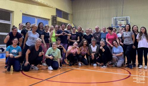 Grupa kobiet w różnym wieku pozuje do wspólnego zdjęcia na sali gimnastycznej, uśmiechając się i wykonując gesty serc lub kciuków uniesionych w górę.
