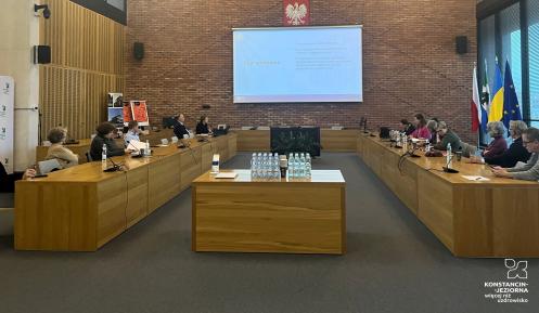 Spotkanie konsultacyjne w sali obrad, podczas którego uczestnicy omawiają strategię rozwoju gminy, a na ekranie wyświetlany jest plan spotkania.