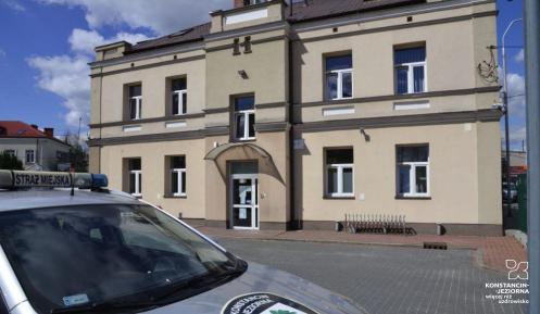 Piętrowy budynek straży miejskiej, na pierwszym planie fragment samochodu z herbem gminy na masce.