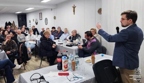 Mężczyzna w marynarce przemawia do zgromadzonych mieszkańców siedzących przy stołach, podczas spotkania w świetlicy wiejskiej.