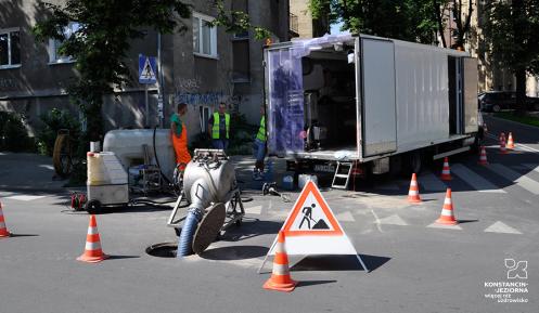 Ekipa pracowników w odzieży ochronnej przeprowadzająca prace związane z konserwacją kanalizacji na miejskiej ulicy, otoczoną pachołkami drogowymi i znakami ostrzegawczymi.