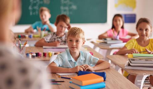 W klasie nauczycielka stoi przy tablicy, dzieci siedzą w ławkach i zgłaszają się do odpowiedzi