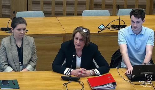 Trzy osoby siedzą przy stole konferencyjnym, a kobieta w środku mówi do mikrofonu, podczas gdy pozostałe osoby słuchają i notują.