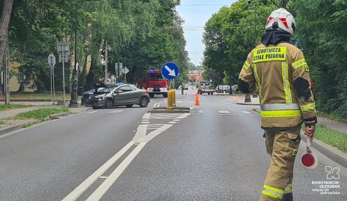 Strażak kieruje ruchem na miejscu wypadku drogowego, gdzie samochód osobowy uderzył w latarnię, a na miejscu obecne są służby ratunkowe.