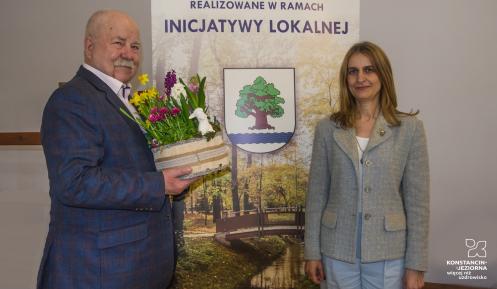 Mężczyzna trzymający kosz z wiosennymi kwiatami i figurką baranka stoi obok kobiety na tle plakatu promującego inicjatywę lokalną.
