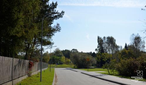 Ulica: po prawej stronie jest chodnik, a po lewej trawnik i płot. Za nim drzewa.